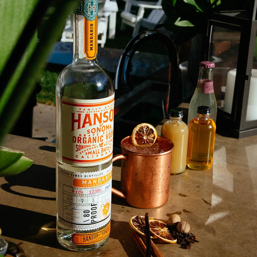 A mulled mule vodka cocktail sat on an outdoor bar in the sunshine, next to a bottle of hanson mandarin vodka