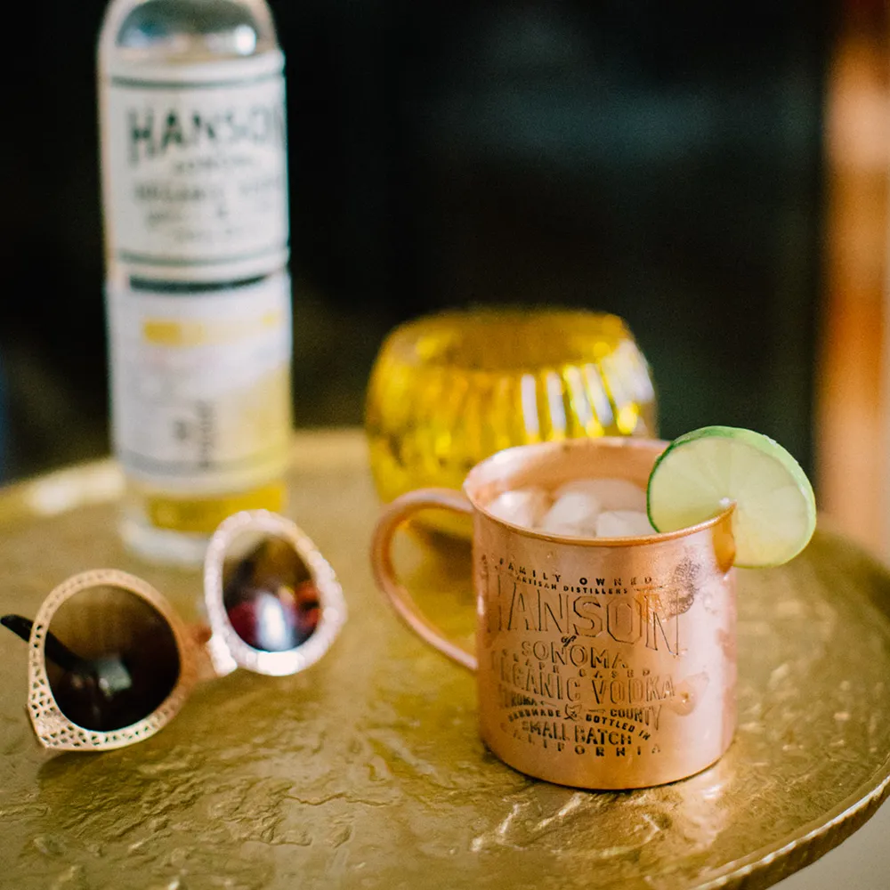 A ginger vodka cocktail in a copper mug on a gold table next to some hanson ginger vodka and some sunglasses